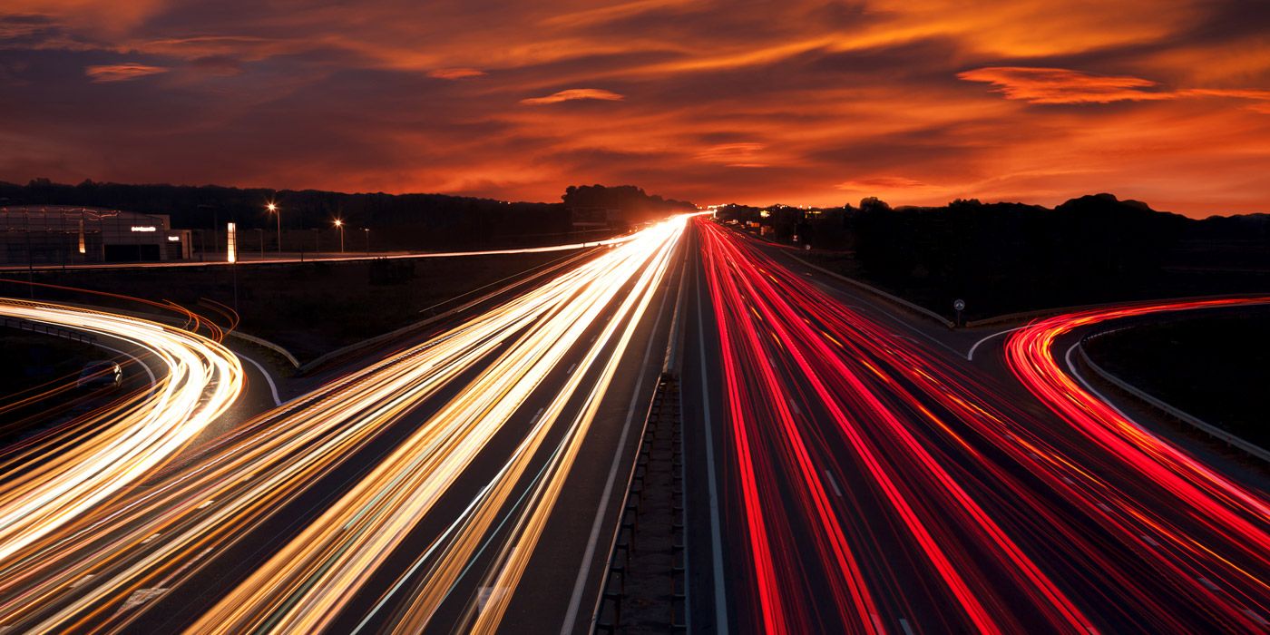 Langzeitbelichtungsfoto einer stark befahrenen Straße in der Dämmerung, links sieht man die weiße Lichterspur der Vorderlichter, rechs die rote der Hinterlichter